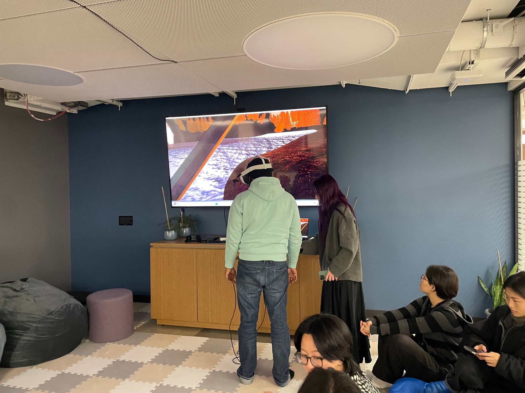 A student in a VR headset demoing their final project at the Renewal Room showcase—their in-headset view mirrored on a large screen behind them, classmates seated on bean bags and cushions watching