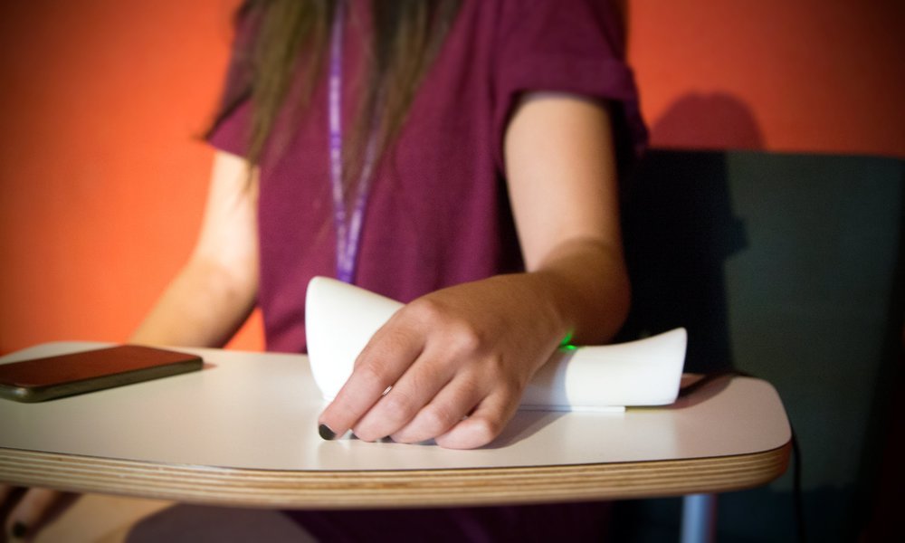 A hand resting on a white device used in the installation — phone nearby, warm orange wall behind
